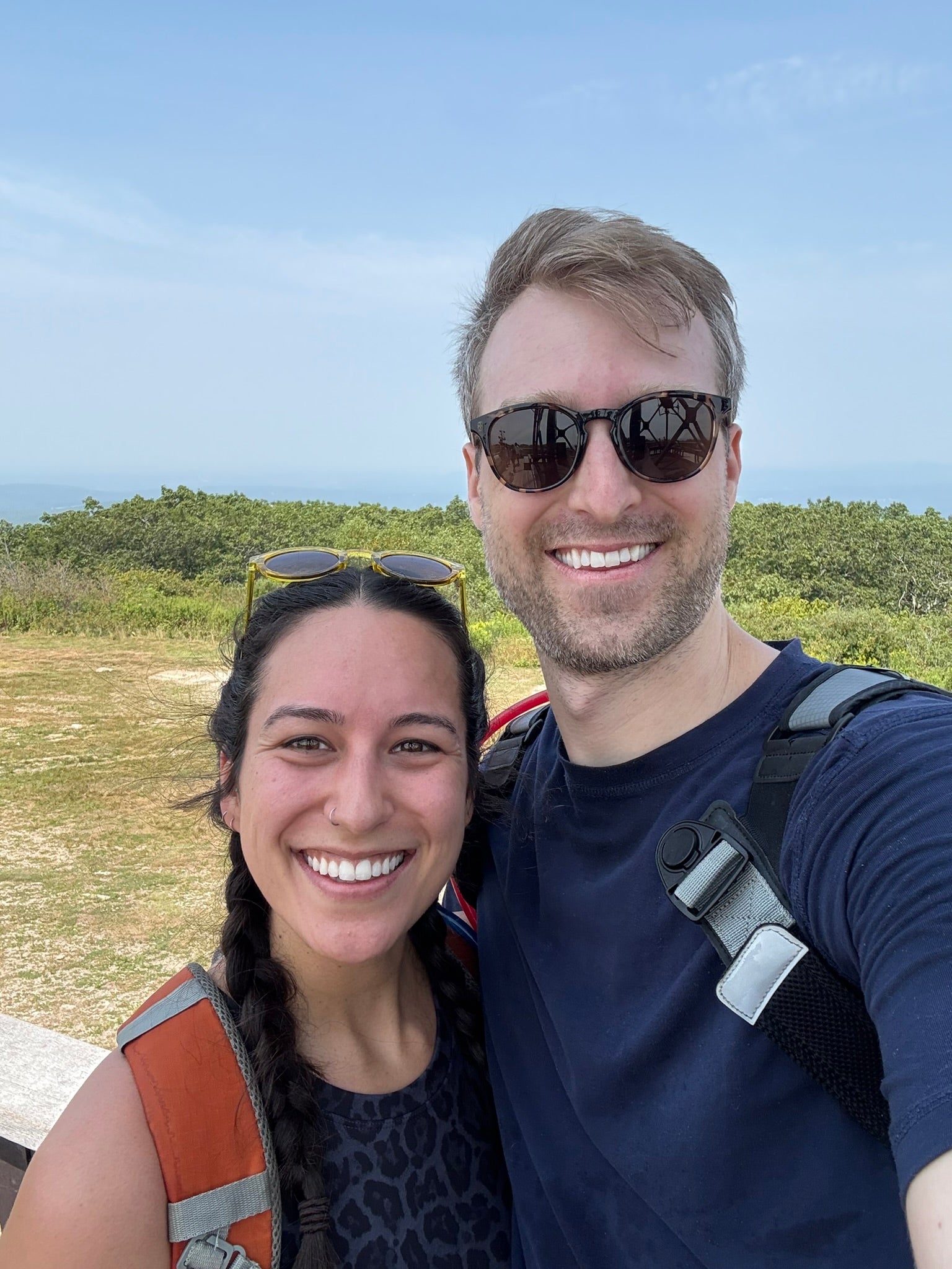 Two people smiling outdoors with backpacks, trees, and open sky